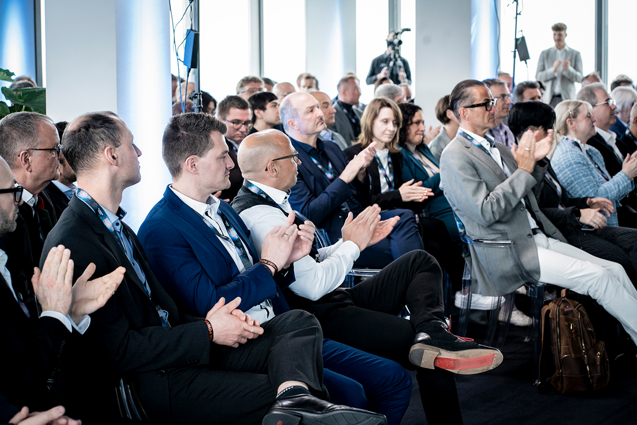 A diverse group of people seated in an event room, clapping together in response to a presentation or performance.