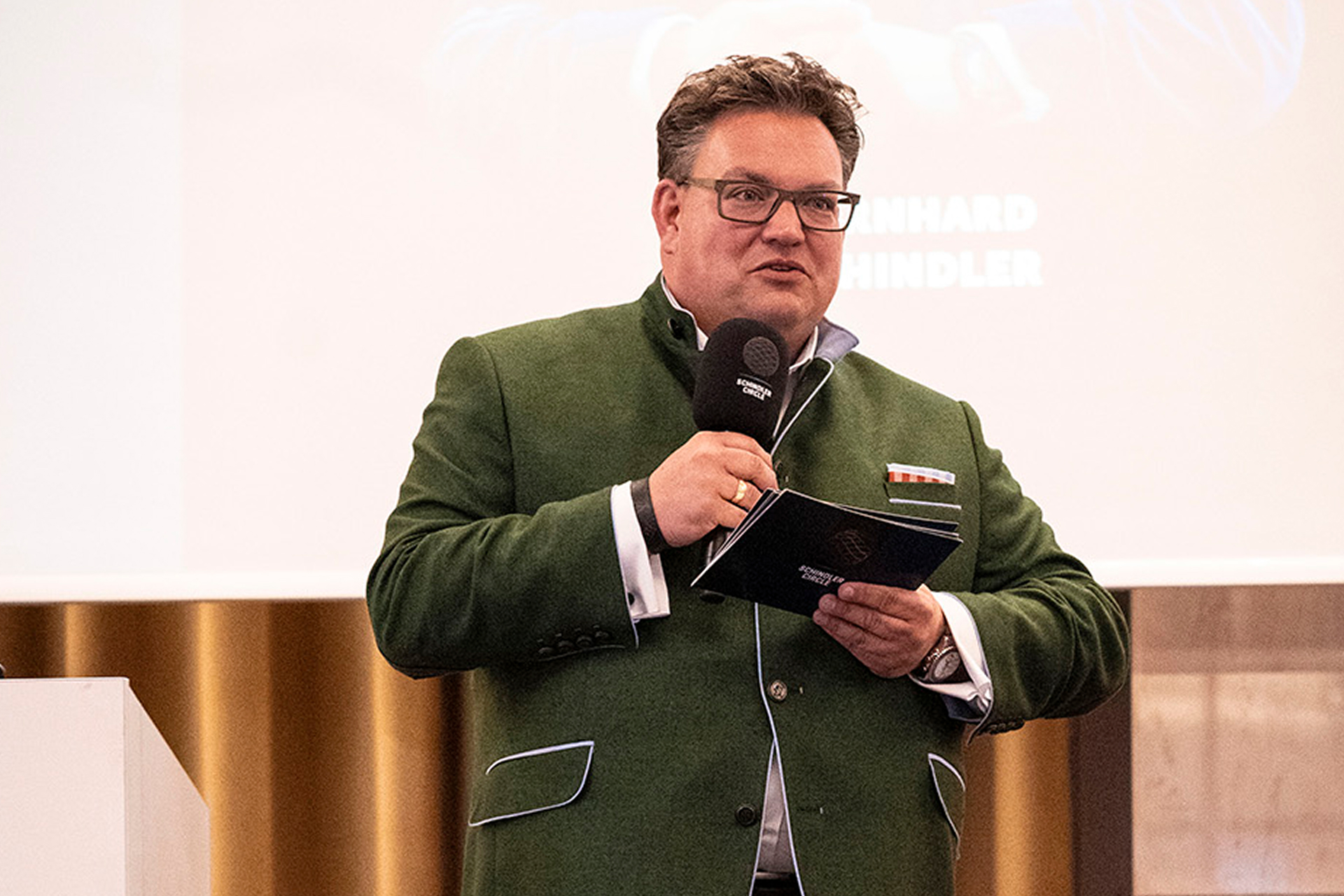A man in a green traditional jacket holds a microphone and booklet while speaking confidently. A presentation screen is in the background.