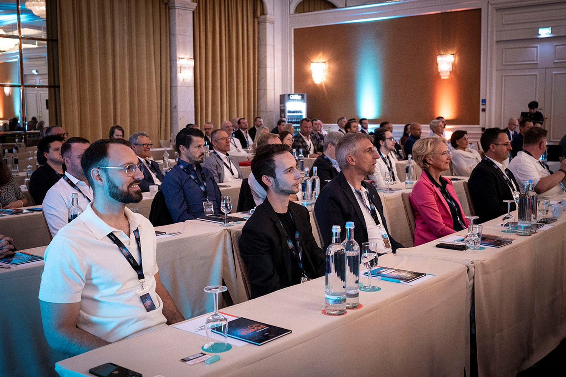 Audience attentively watching a presentation in a conference room with beige curtains. Attendees in business attire sit at tables with water bottles.